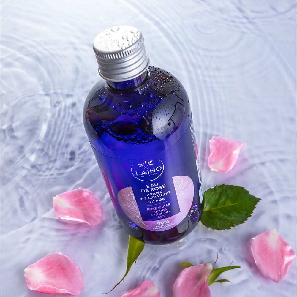 Blauwe fles met zilveren dop in water met rozenblaadjes. Opschrift: LAINO, Eau de Rose, verzacht & verfrist het gezicht.