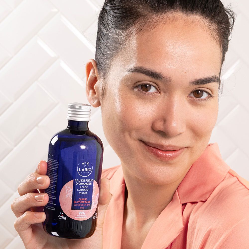 Femme tenant un flacon bleu en verre, bouchon argenté, LAINO Eau de Fleur d'Oranger. Texte: Apaise & adoucit visage. La femme sourit.