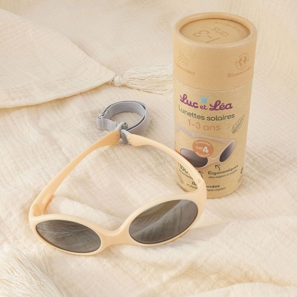 Beige zonnebril met grijze glazen en grijze band. Daarnaast de verpakking. Opschrift: Luc & Léa, zonnebril 1-3 jaar.