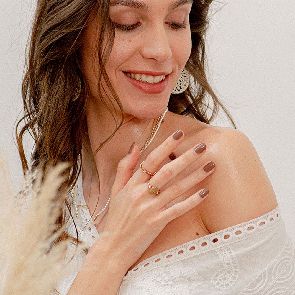 Vrouw met bruin haar, draagt sieraden. Ze raakt haar schouder aan. Lichte kleding.