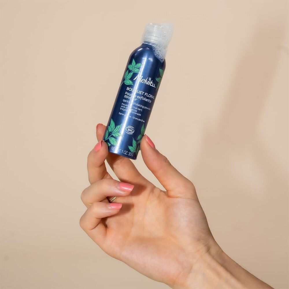Hand houdt blauwe fles met witte dop. Opschrift: Melvita, Bouquet Floral, Poudre exfoliante douceur. Bio-certificering.