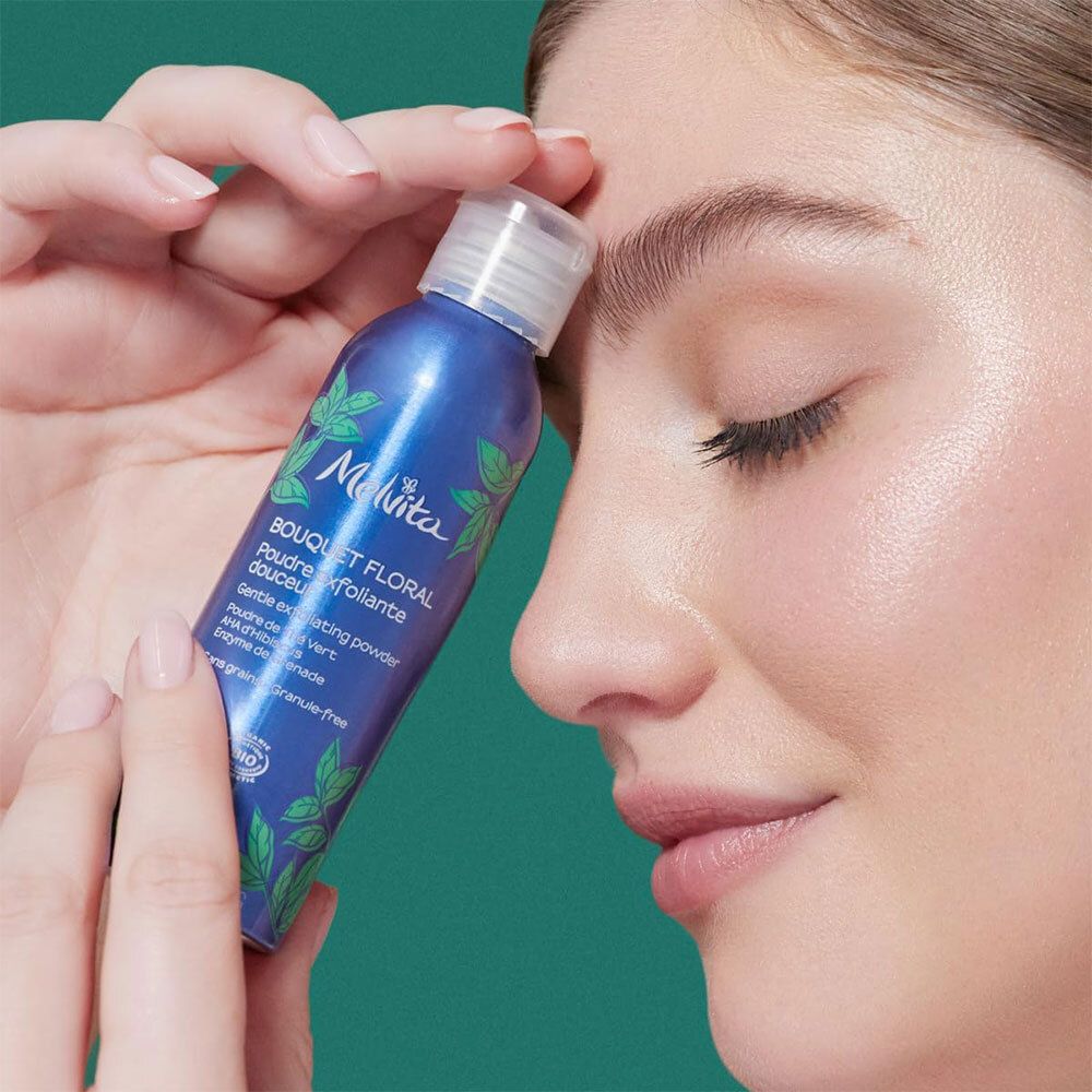 Melvita peelingpoeder fles in een wolk van wit poeder. Blauwe fles met groene bladeren.