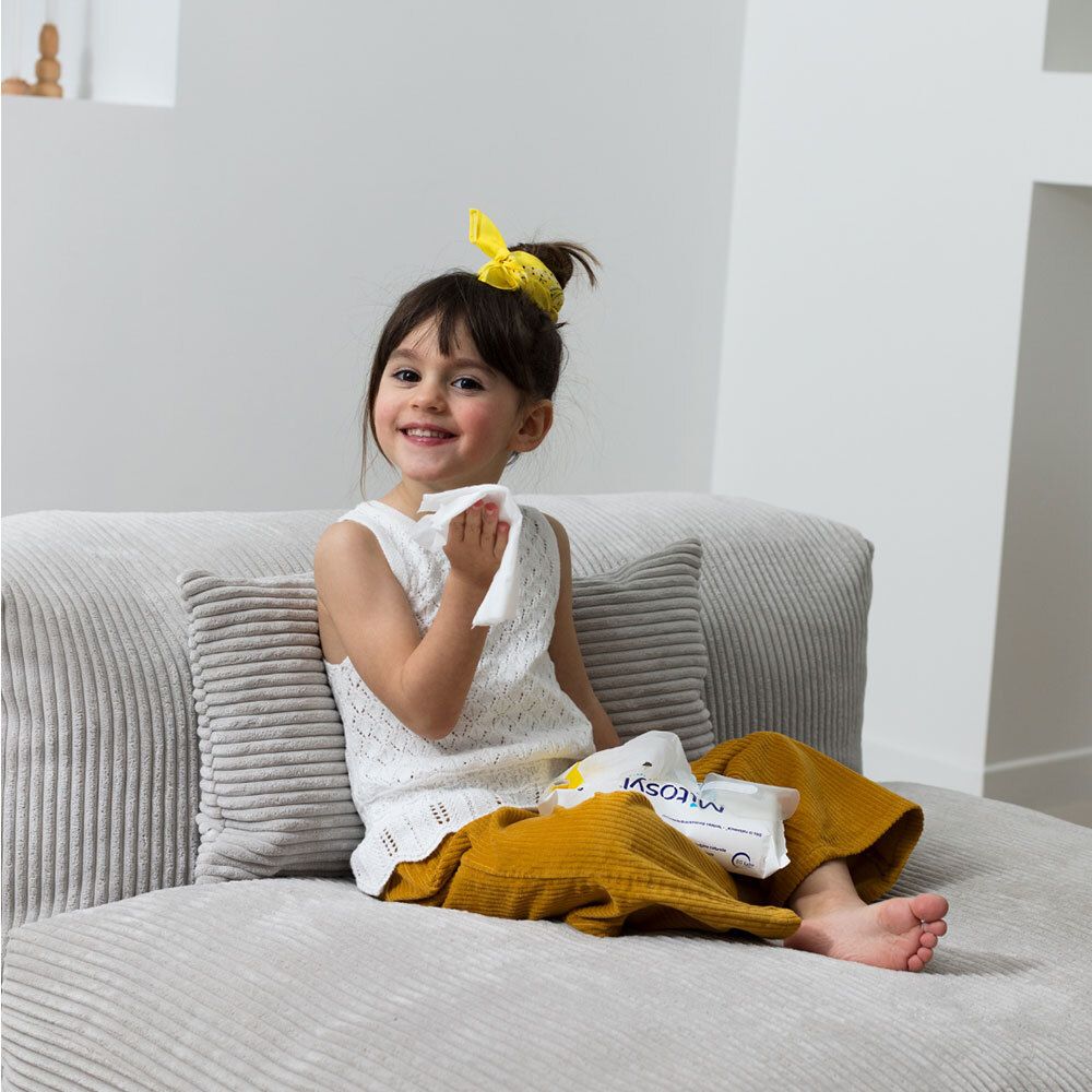 Enfant assise sur un canapé tenant une lingette. Un paquet "Mitosyl Lingettes à l'eau" est à côté.
