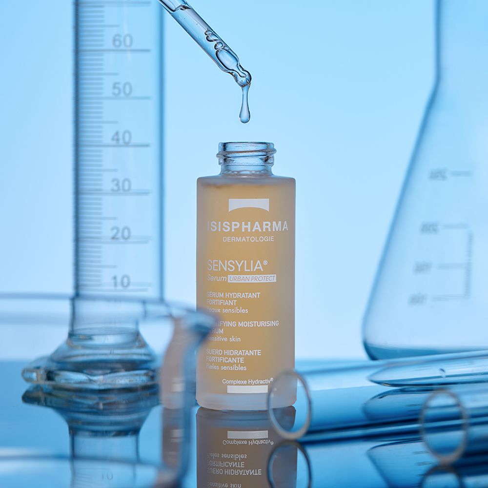 Flacon de sérum avec pipette, gouttes tombant dedans. Matériel de laboratoire en arrière-plan. ISISPHARMA Sensylia.