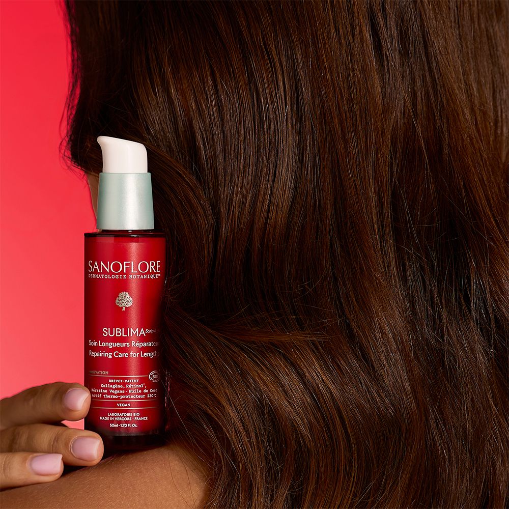 Flacon rouge avec pompe blanche. Inscription: SANOFLORE, SUBLIMA. Le flacon est tenu sur les cheveux.