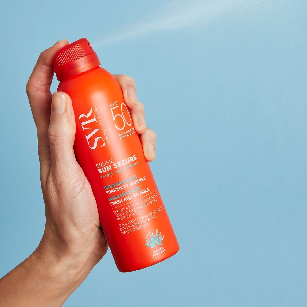 Een hand houdt een oranje spray. Spuitnevel zichtbaar. Opschrift: SVR, SPF 50, Sun Secure Brume. Blauwe achtergrond.