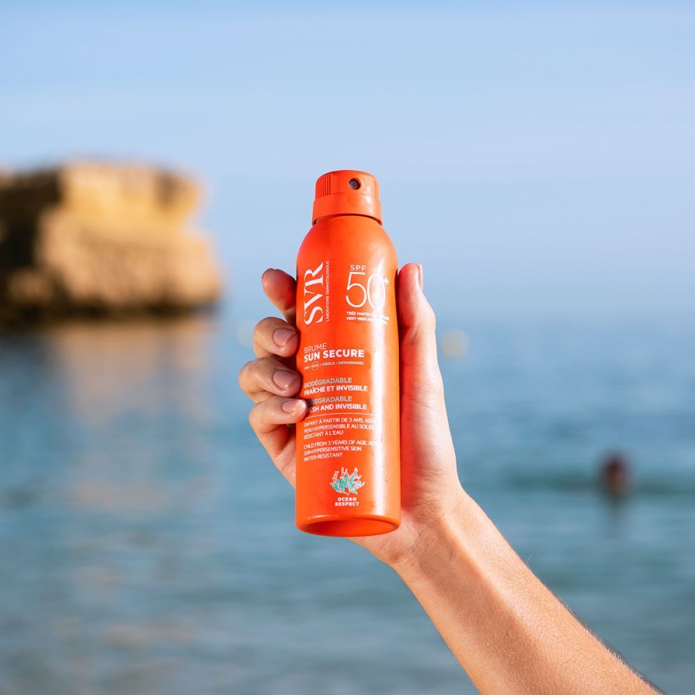 Hand houdt een oranje spray op het strand. Opschrift: SVR, SPF 50, Sun Secure Brume. Zee op de achtergrond.
