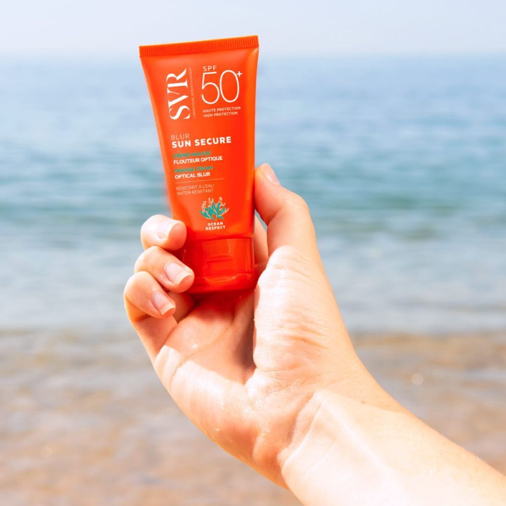 Hand houdt een tube SVR Sun Secure Blur LSF50+ vast. Oranje tube met witte tekst. Zee op de achtergrond.