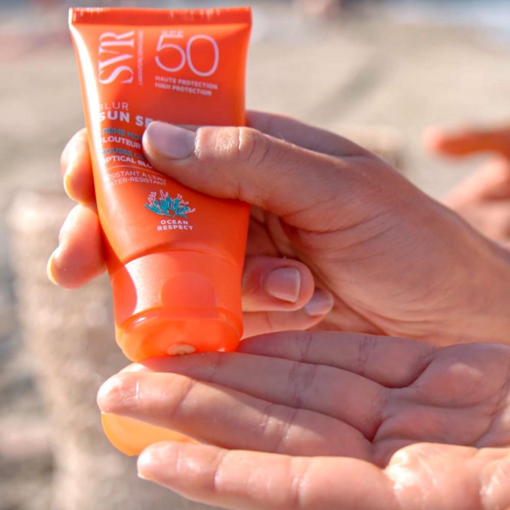 Hand houdt een tube SVR Sun Secure Blur LSF50+ vast. Oranje tube met witte tekst. Zee op de achtergrond.