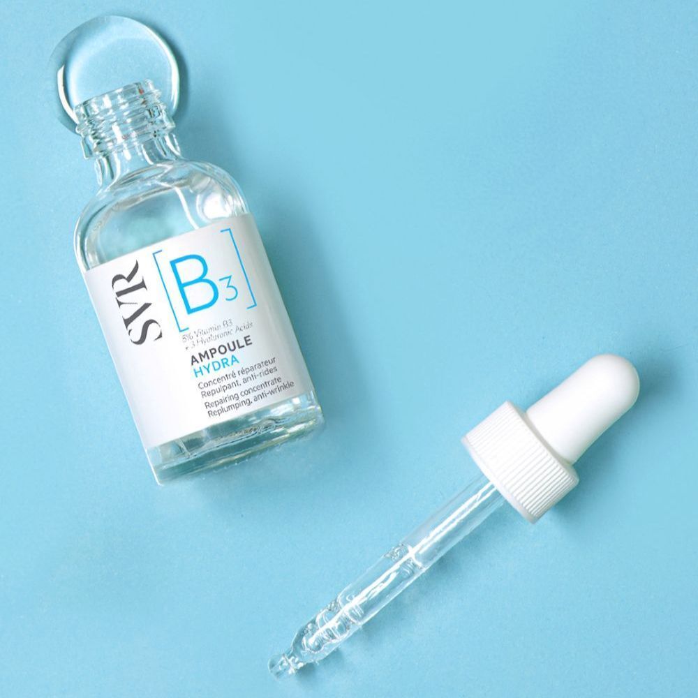 Een persoon houdt een fles SVR [B3] Ampulle Hydra vast. Blauwe tegels op de achtergrond.