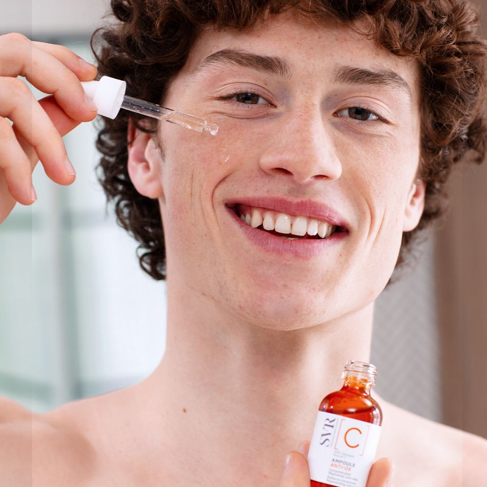 Man houdt flesje met druppelaar. Druppels op gezicht. Etiket: SVR [C] Ampoule Anti-Ox.