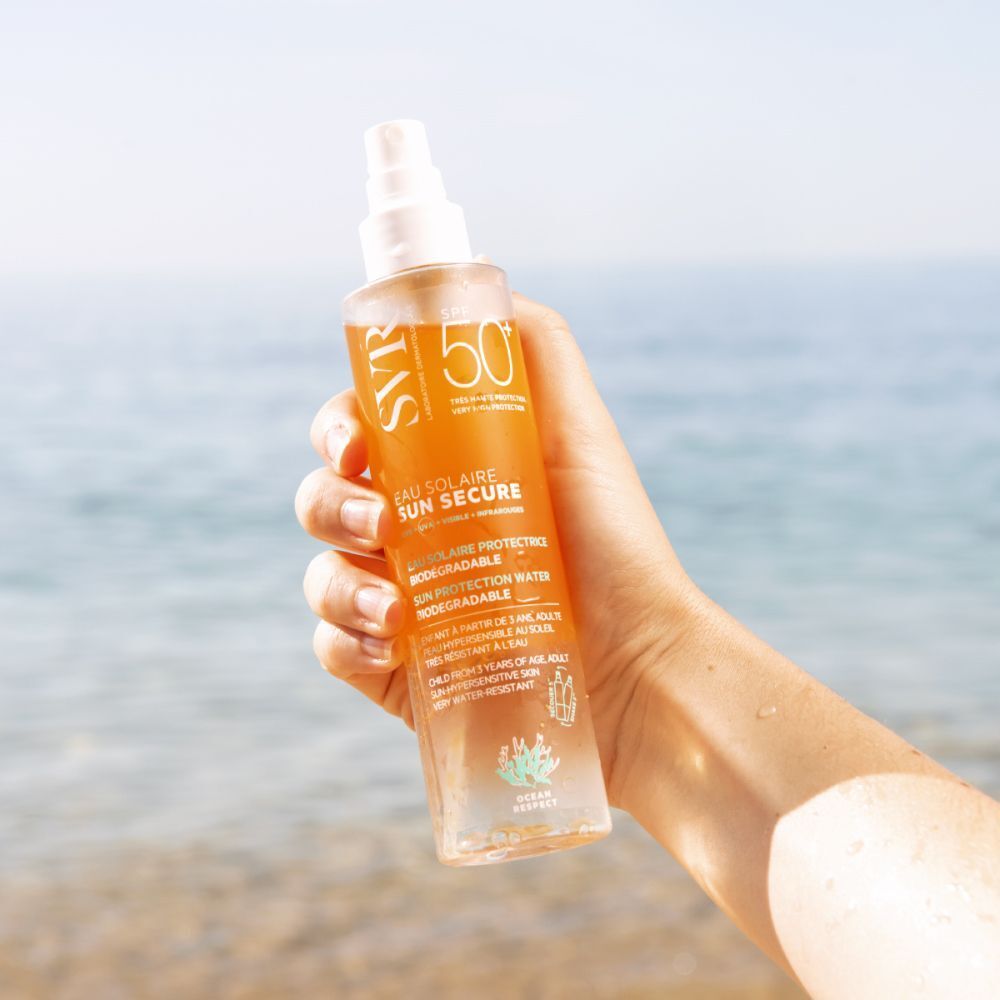 Une main tient un flacon de SVR SUN SECURE Eau de Solaire LSF 50+. Étiquette jaune, tête de pulvérisation blanche. Fond : mer.