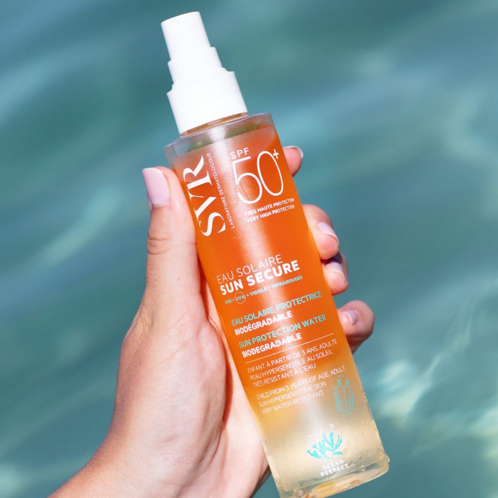 Une main tient un flacon de SVR SUN SECURE Eau de Solaire LSF 50+. Étiquette jaune, tête de pulvérisation blanche. Fond : eau.