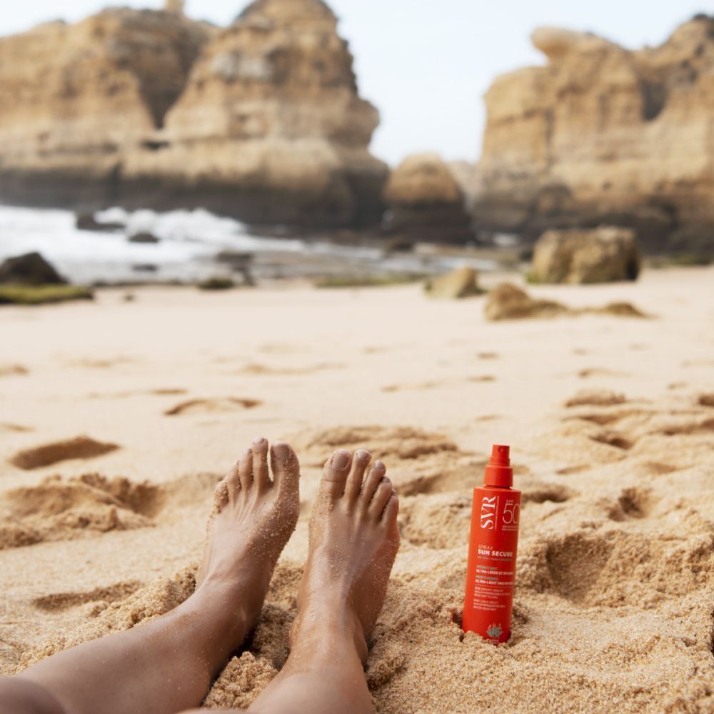 Een oranje fles ligt op het strand naast voeten. Opschrift: SVR, SUN SECURE, SPF 50+.