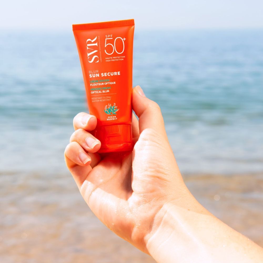 Main tenant un tube de crème SVR Sun Secure SPF50+. Tube orange avec texte blanc. Texte: Blur, Sun Secure, Creme Mousse, Flouteur Optique, Optical Blur.