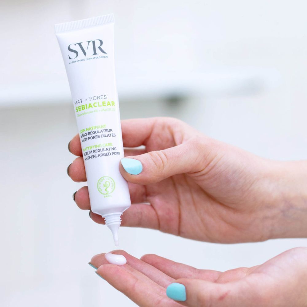 Tube SVR Sebiaclear Matte + Poren wordt in één hand gedrukt. Witte crème wordt in de andere hand gedrukt. Lichte huid, blauwe nagels.