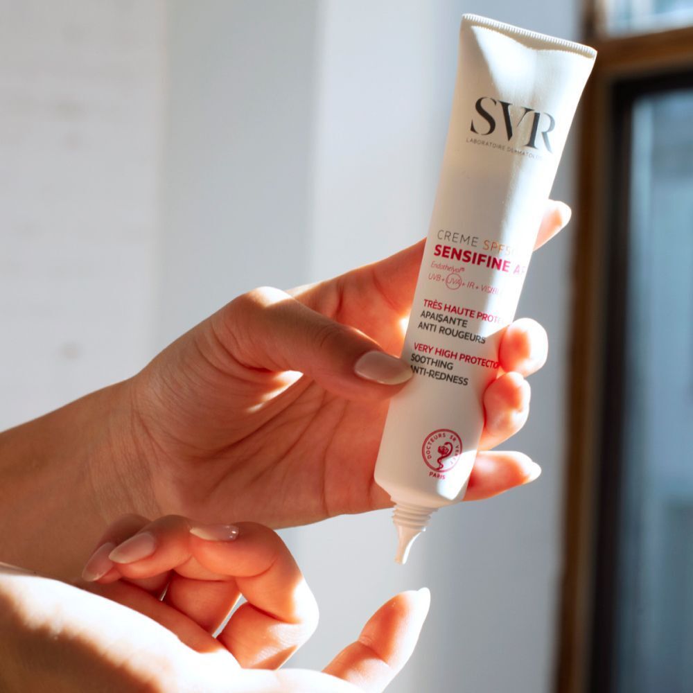 Un tube de crème SVR Sensifine AR LSF50. Tube blanc avec texte rouge et noir. Inscription: Crème SPF50+ Sensifine AR. Logo: SVR. Tenue par des mains.