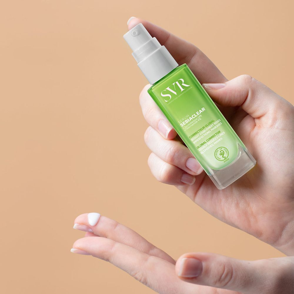Une main tient un flacon de sérum SVR SEBIACLEAR vert. Inscription : SEBIACLEAR, anti-imperfections, anti-rides.