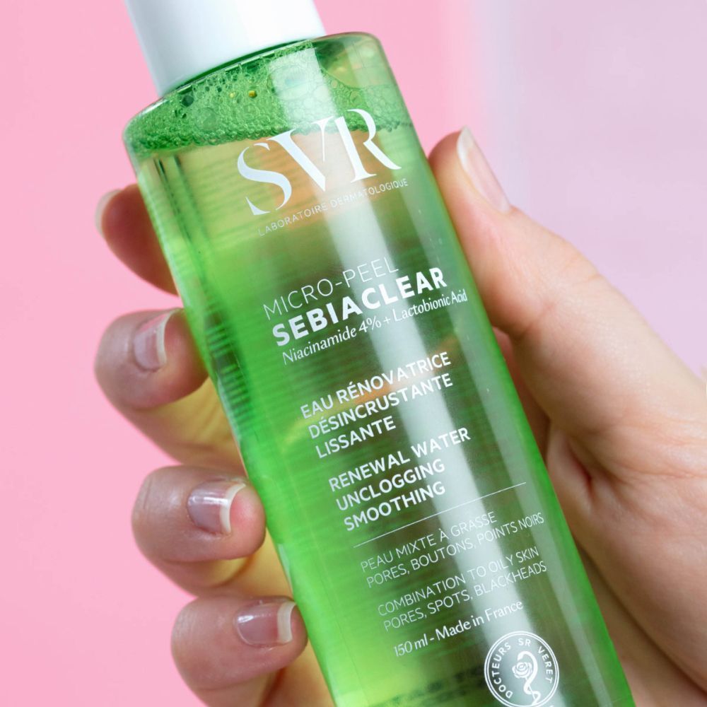 Groene fles met witte dop. Opschrift SVR SEBIACLEAR Micro-Peel. Tekst: Eau rénovatrice désincrustante lissante. Hand houdt fles vast.