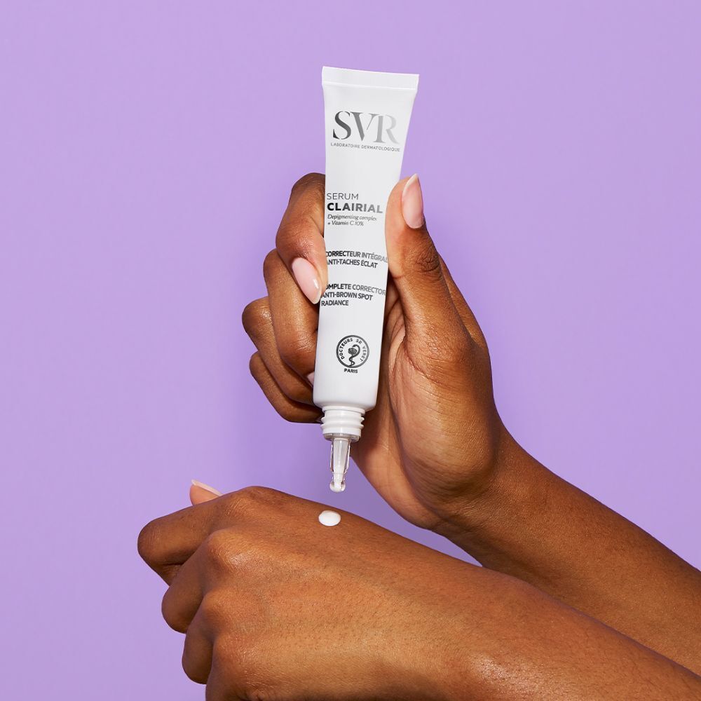 Handen met SVR Clairial Serum. Tube wordt op de hand aangebracht. Paarse achtergrond. Tekst: Clairial Serum, Corrector intégral anti-taches éclat.