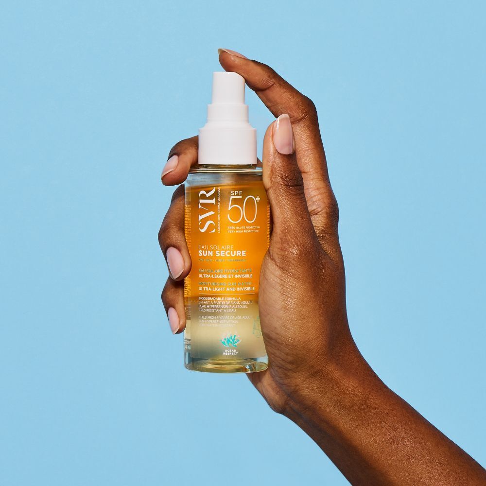 Hand houdt een fles SVR Sun Secure Eau Solaire SPF50+. Gele vloeistof, witte spraykop. Tekst: SPF50+, Eau Solaire. Blauwe achtergrond.