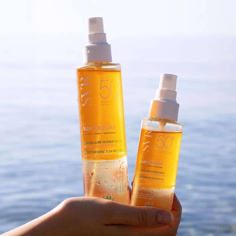 Hand houdt twee flessen SVR Sun Secure Eau Solaire SPF50+. Gele vloeistof, witte spraykop. Tekst: SPF50+, Eau Solaire Hydratante. Zee op de achtergrond.
