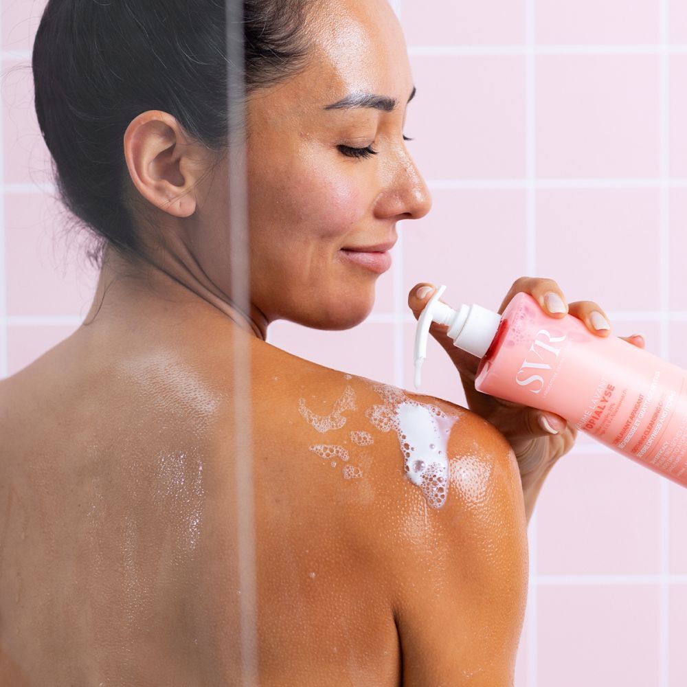 Vrouw onder de douche, achteraanzicht. Ze houdt een roze fles met witte pomp vast. Schuim op de schouder.