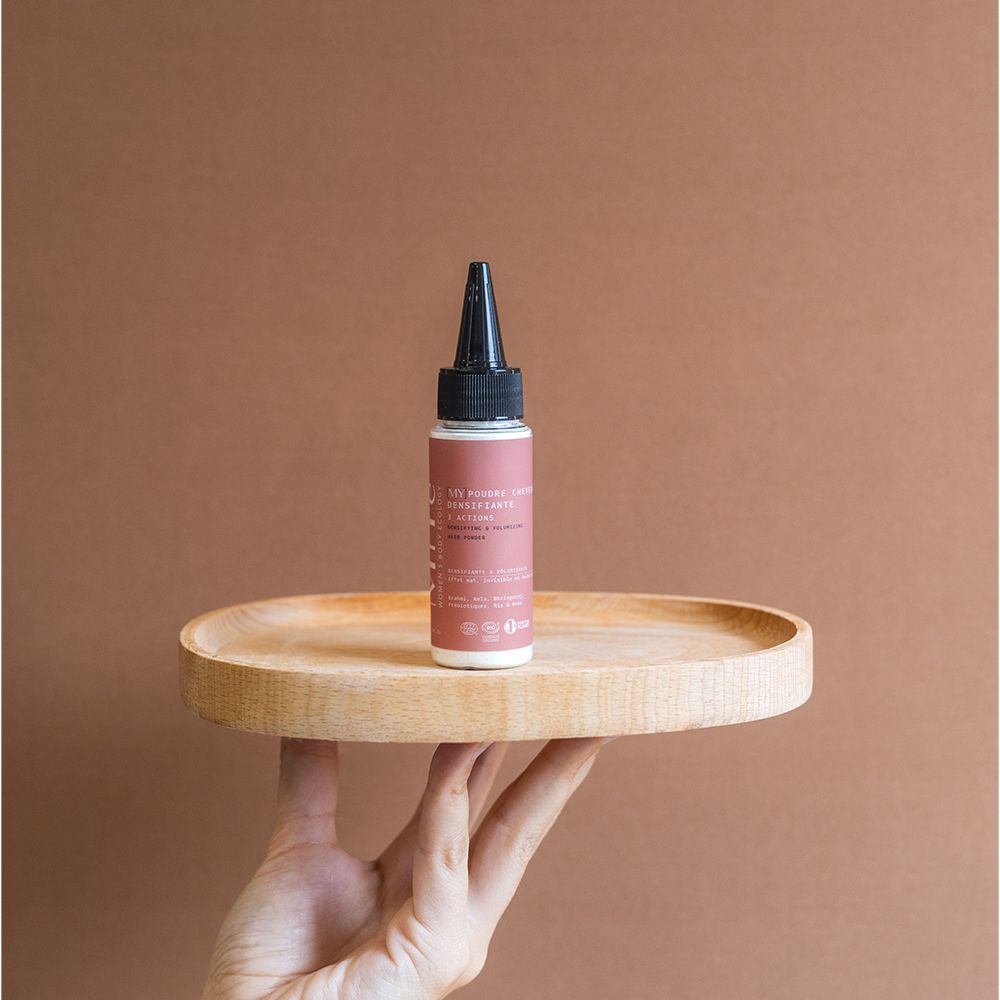 Une petite bouteille plate de couleur rose avec un embout applicateur noir repose sur un plateau rond en bois.