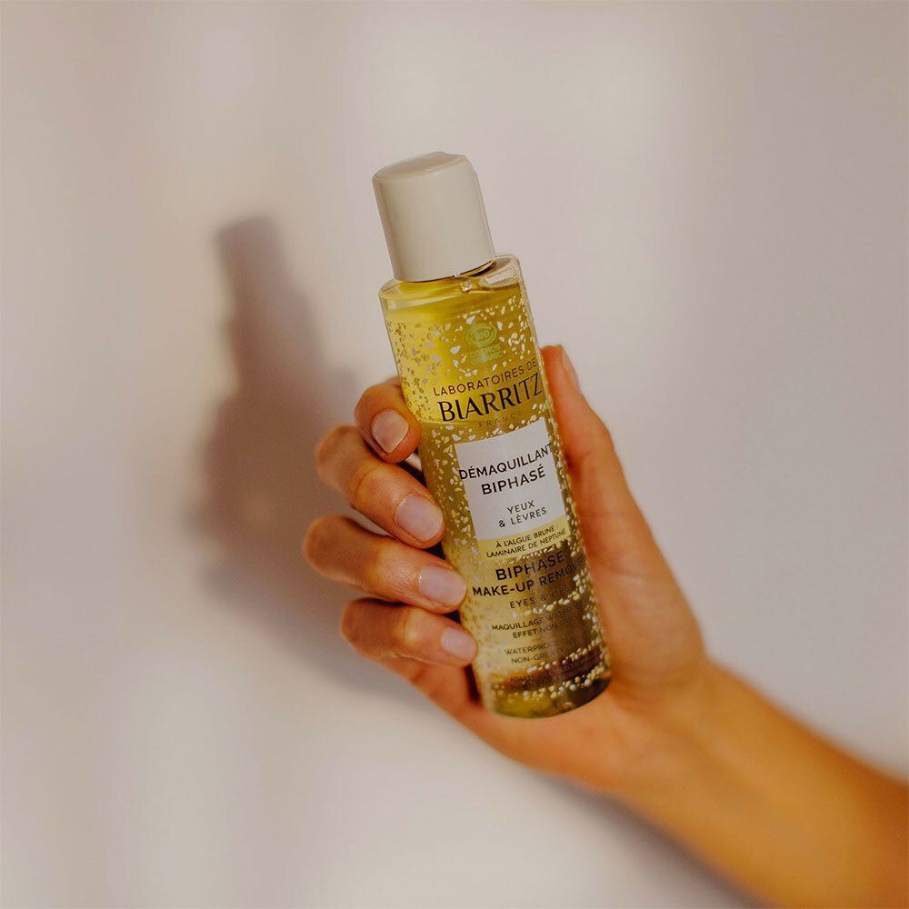 Een hand houdt een fles Démaquillant Biphasé Yeux & Lèvres Bio vast. De achtergrond is wit.
