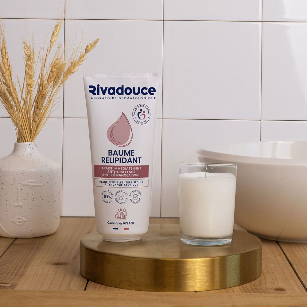 Product op gouden oppervlak. Witte tube met Rivadouce-logo. Kaars en vaas met graanaren op de achtergrond. Tegelwand.