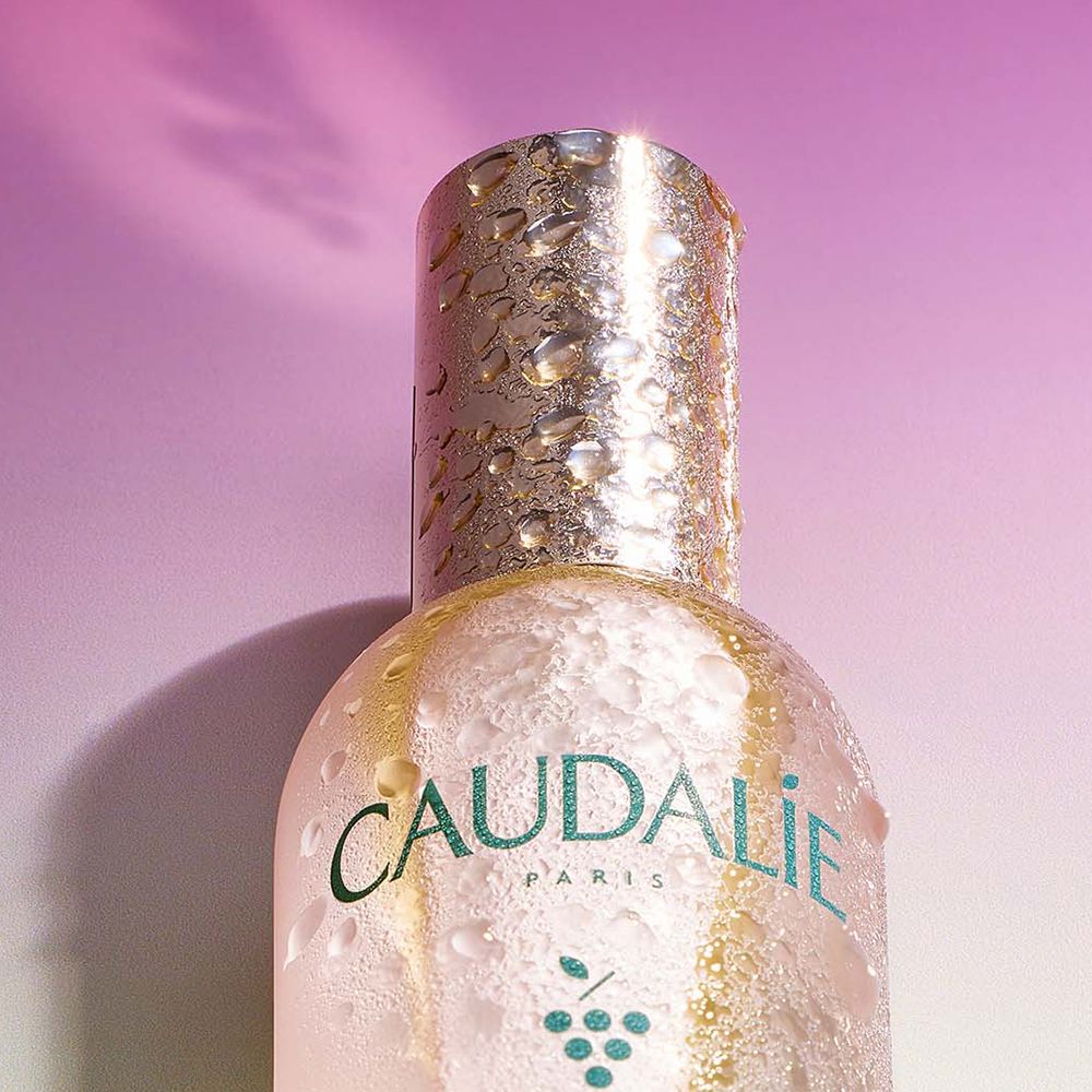 Main vaporisant Caudalie Eau de Beauté. Flacon avec bouchon argenté. Texte: Caudalie, Beauty Elixir.
