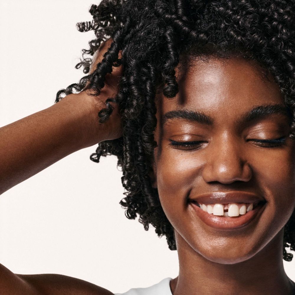 Vrouw met krullend haar. Ze lacht en raakt haar haar aan.