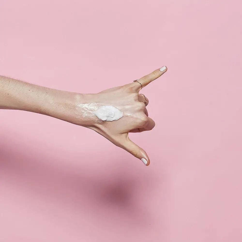 Hand met product op de rug van de hand. Achtergrond roze. Geen verdere details.