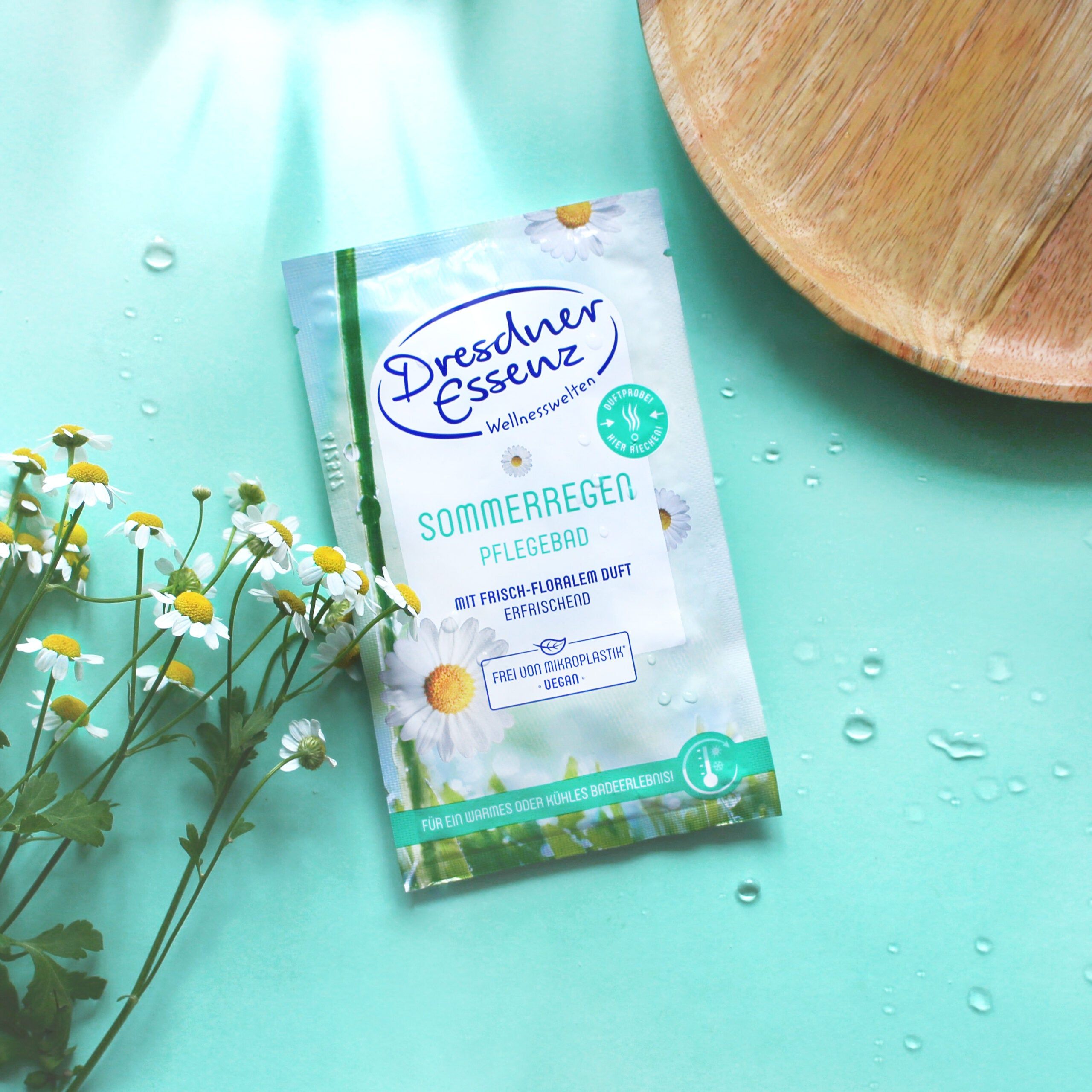 Sachet Dresdner Essenz Pflegebad Sommerregen, fleurs de camomille et bol en bois sur fond bleu.