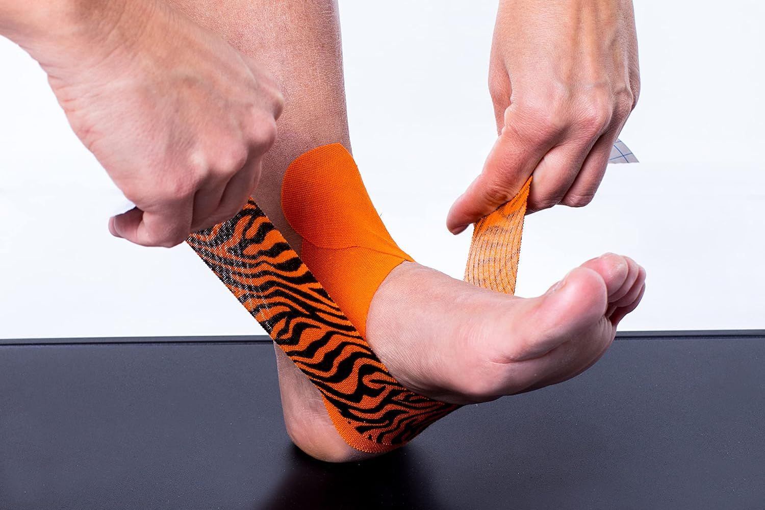Aanbrengen van CureTape kinesiologie tape op de voet. Handen brengen de oranje-zwarte tijgerprint tape aan.