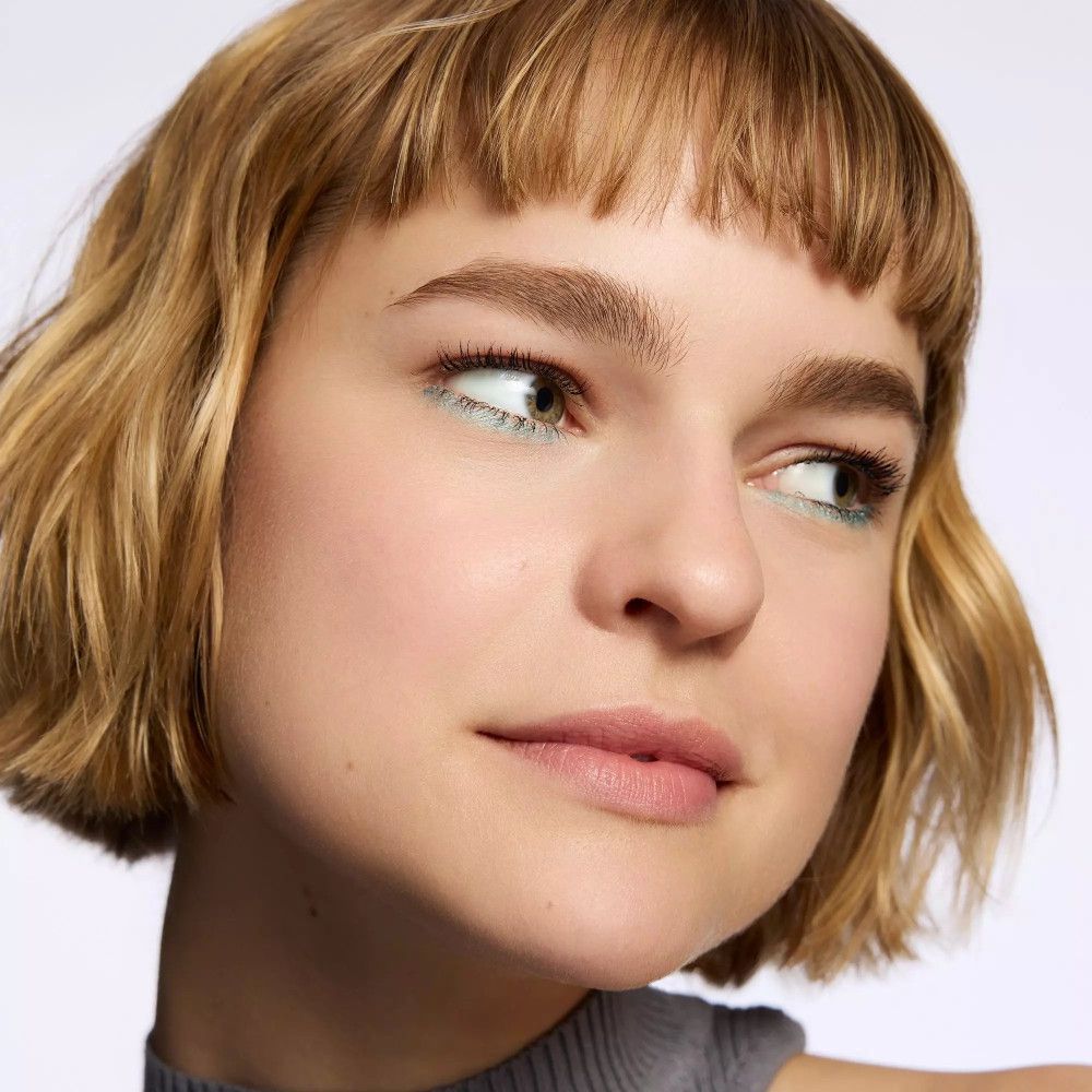 Gros plan d'une femme avec une ligne d'ombre à paupières bleu clair. Cheveux blonds courts, haut gris. Regard de côté.