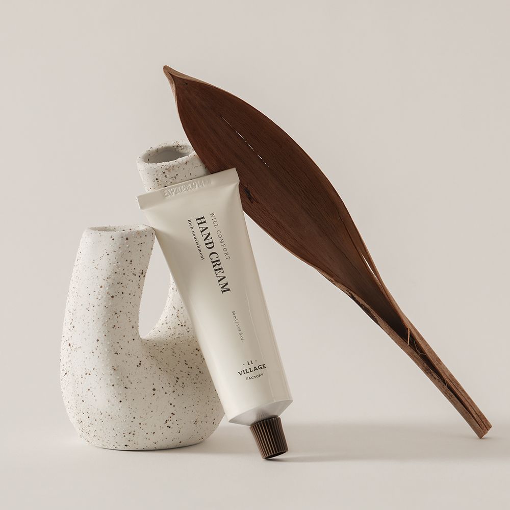 Crèmetube met tekst HAND CREAM, VILLAGE FACTORY, naast een vaas en een blad. Beige tube.