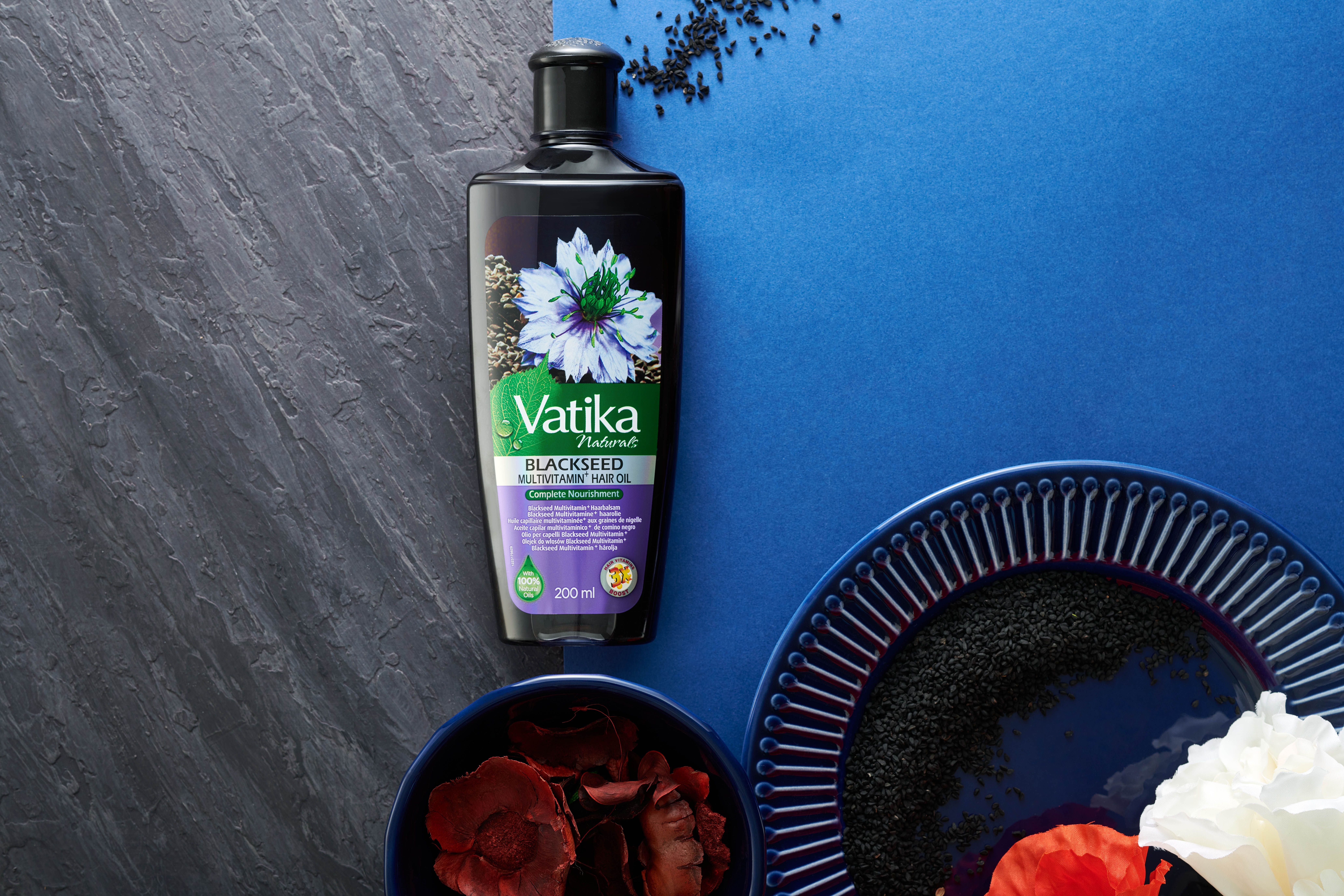 Vatika Naturals Schwarzkümmel haarolie op blauwe achtergrond. Naast kommen met ingrediënten en een bord.