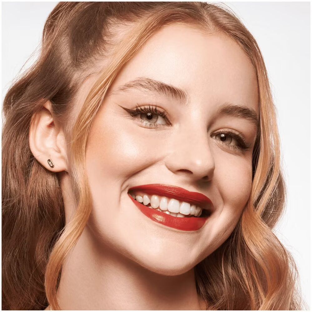 Gros plan d'un visage souriant maquillé. Rouge à lèvres rouge, eye-liner, fard à joues. Cheveux blonds.