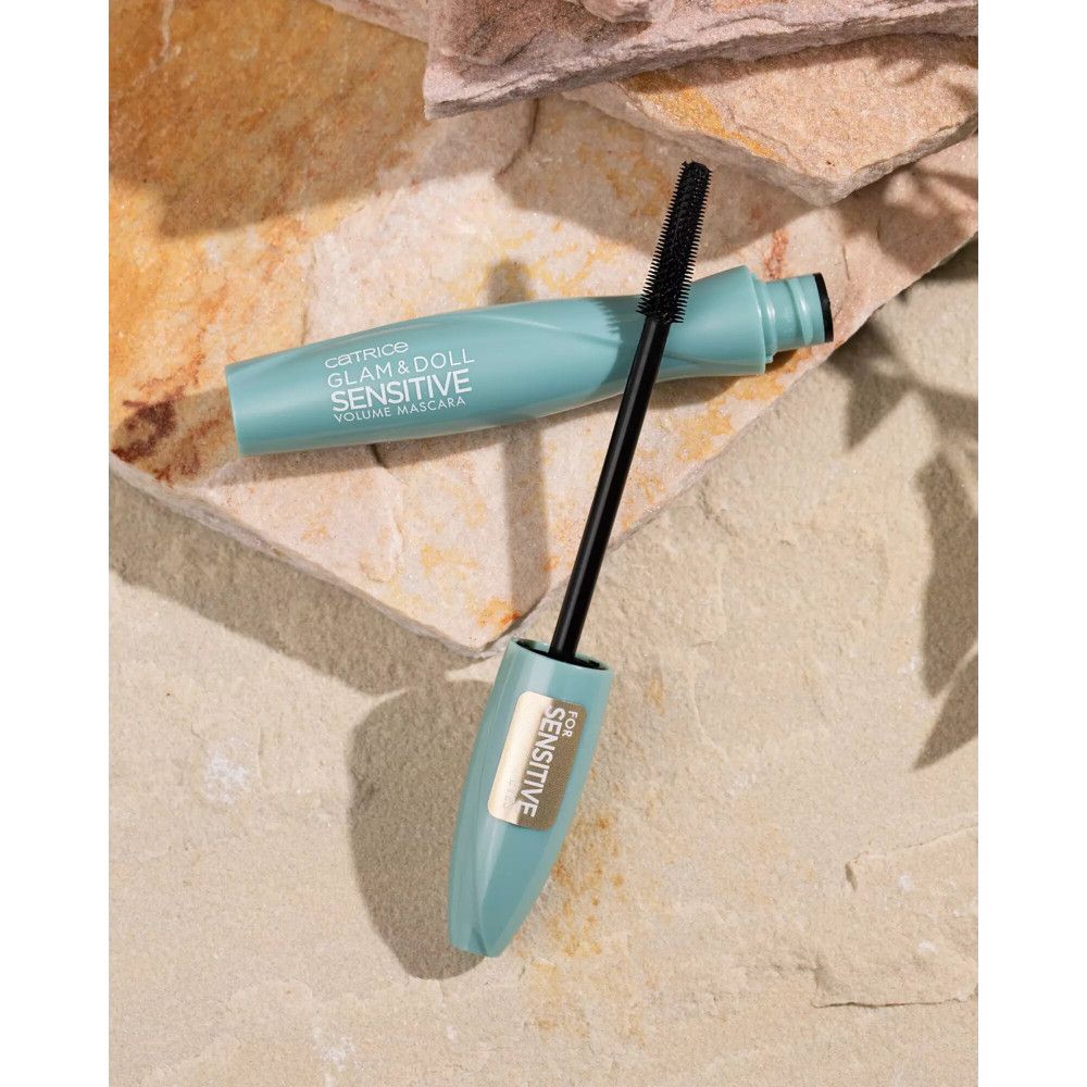 Tube et brosse de mascara turquoise sur fond de pierre. Inscription : Glam & Doll Sensitiv. Brosse sortie.