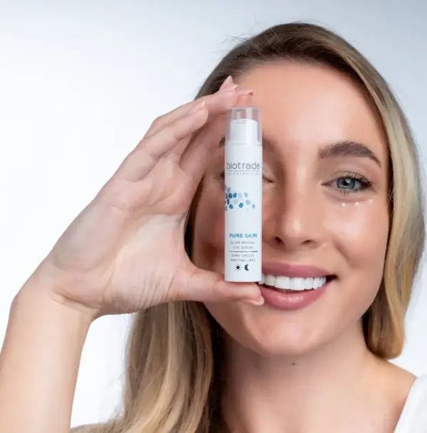 Femme tenant un flacon de crème pour les yeux Biotrade Pure Skin. Flacon blanc avec motif bleu. Texte : Pure Skin Glow Revival Eye Serum.