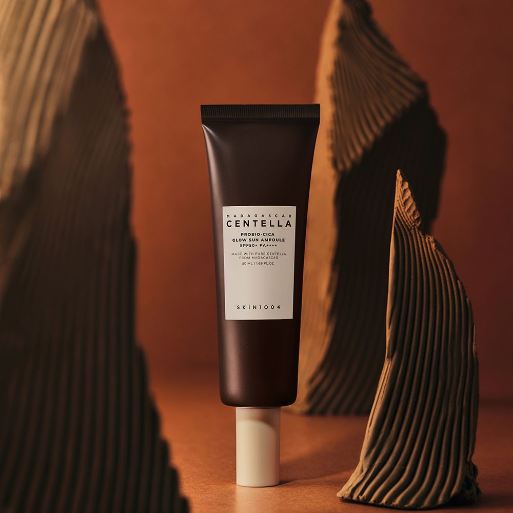 Tube marron avec étiquette blanche. Texte: MADAGASCAR CENTELLA, PROBIO-CICA GLOW SUN AMPULLE SPF50+ PA++++. Sur socle.