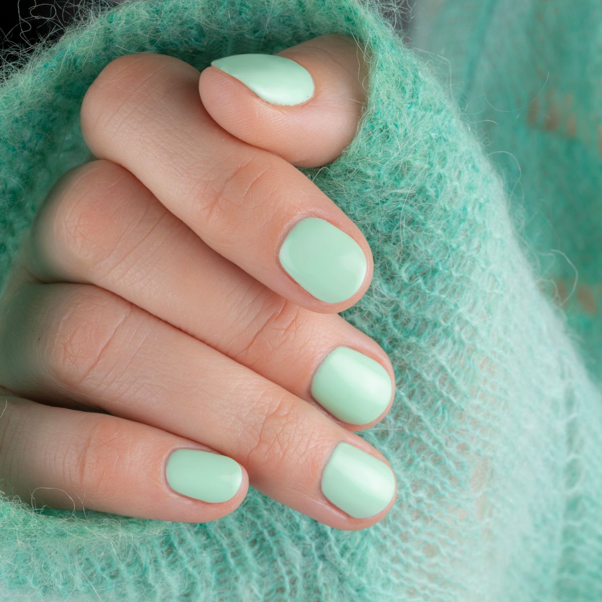 Main avec ongles vernis vert menthe. Les ongles sont courts et de forme ovale. La main est enveloppée dans un pull vert.