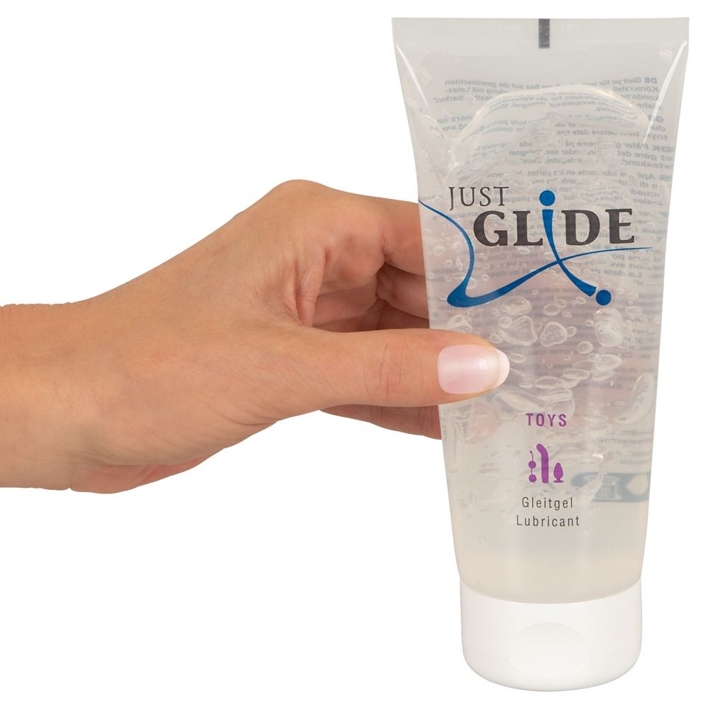 Een hand houdt een tube glijmiddel vast. Opschrift: JUST GLIDE, TOYS, Gleitgel, Lubricant. Transparante gel zichtbaar.