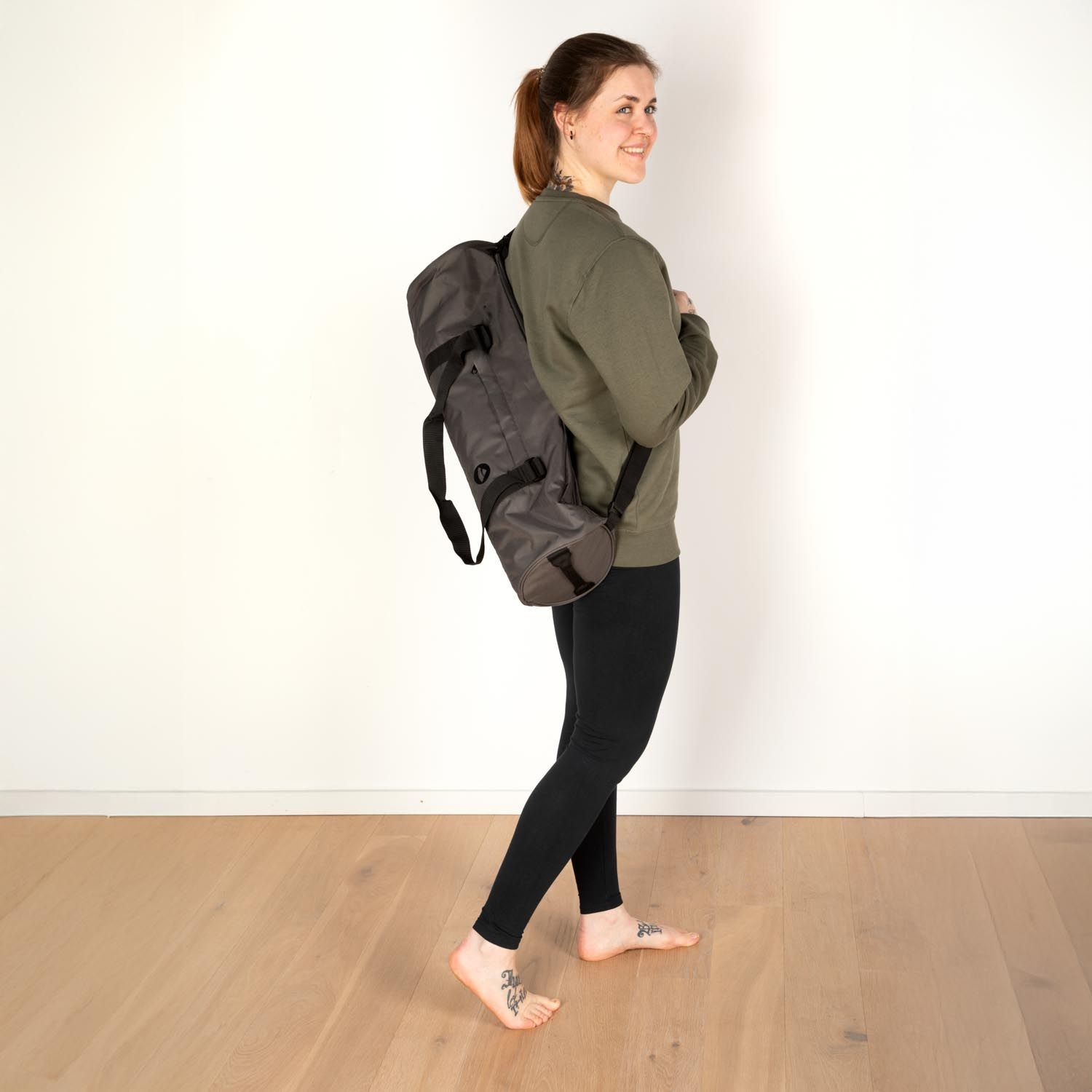 Femme portant un sac de sport gris sur le dos. Legging noir, haut vert. Plancher en bois.