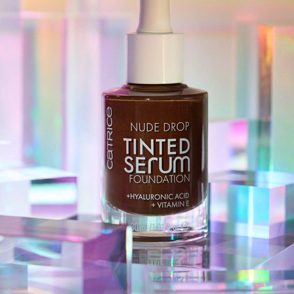 Flacon marron avec bouchon et pipette blancs. Inscription : Nude Drop Tinted Serum Foundation, + Acide hyaluronique + Vitamine E. Sur fond brillant.