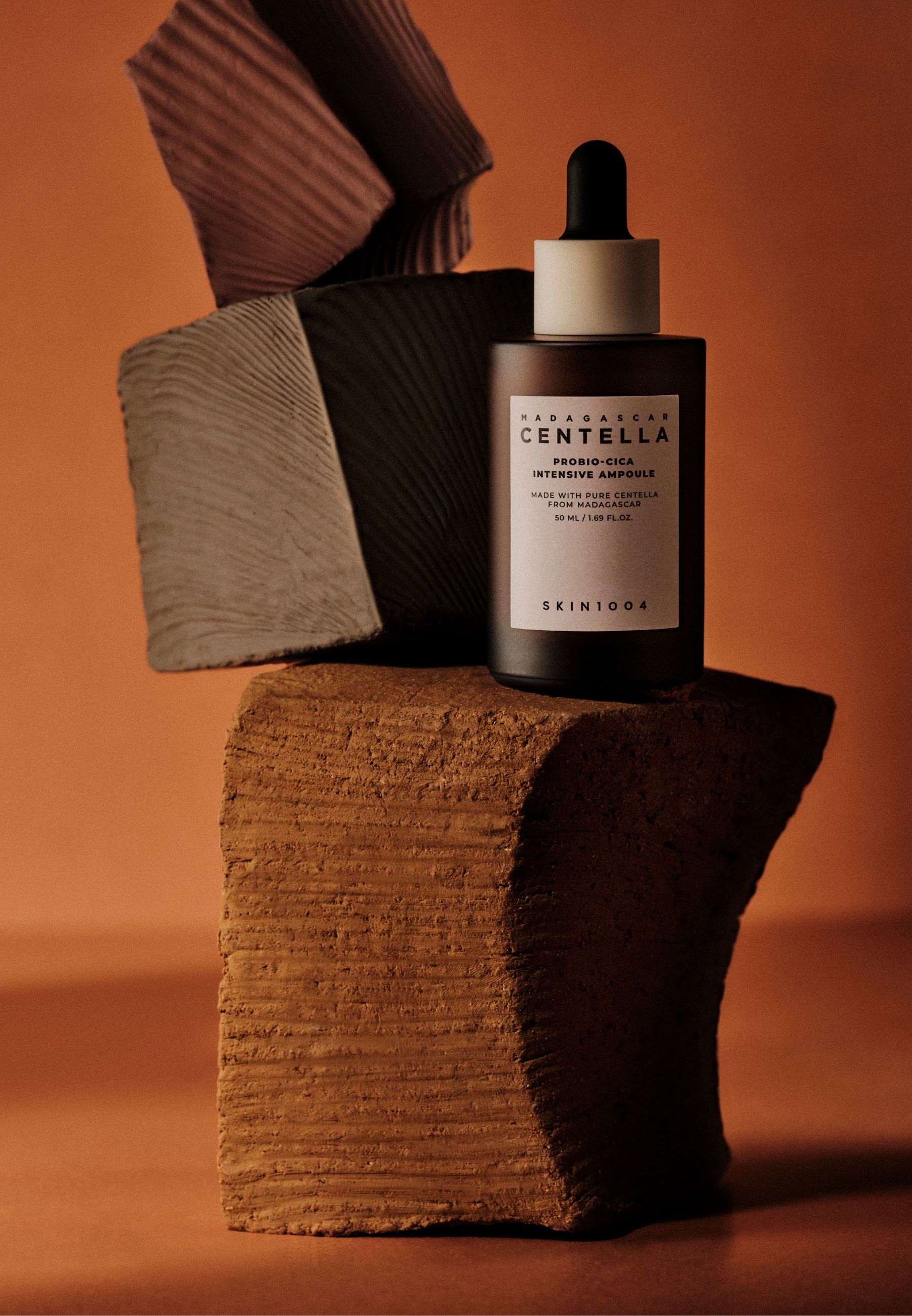 Bruine fles met pipet op stenen. Tekst: CENTELLA, PROBIO-CICA, SKIN1004. Achtergrond: oranje.