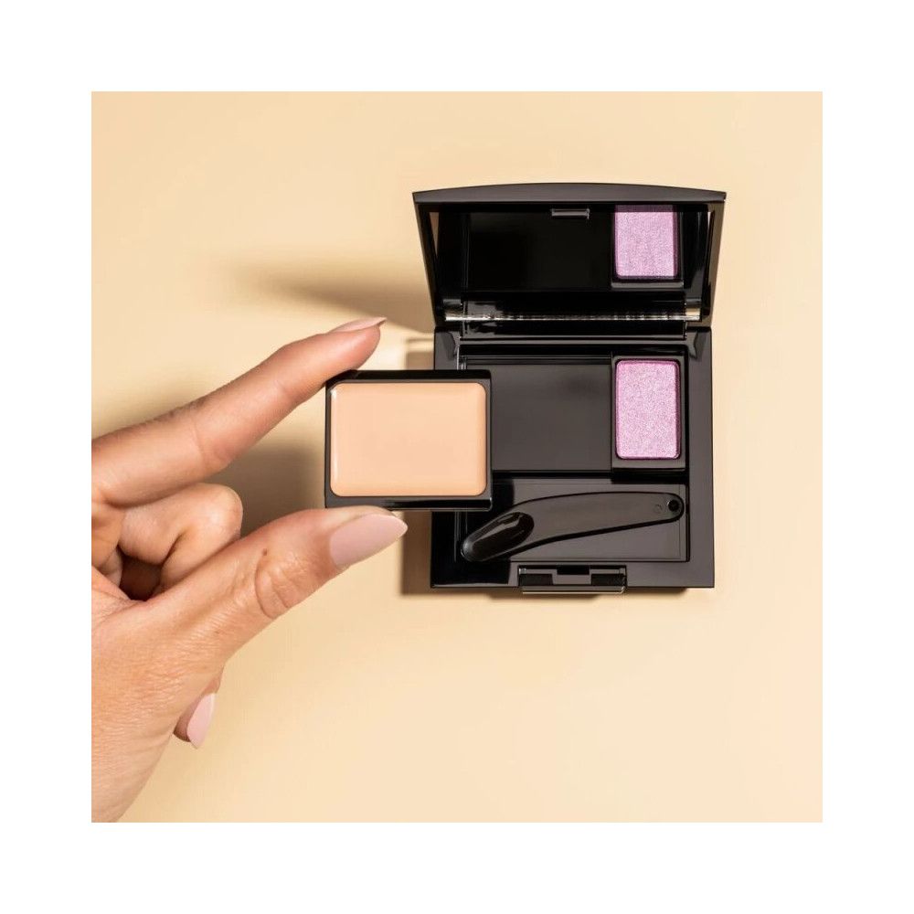 Boîte à beauté Trio ouverte avec fards à paupières et applicateur. Une main tient un autre fard. Fond beige.