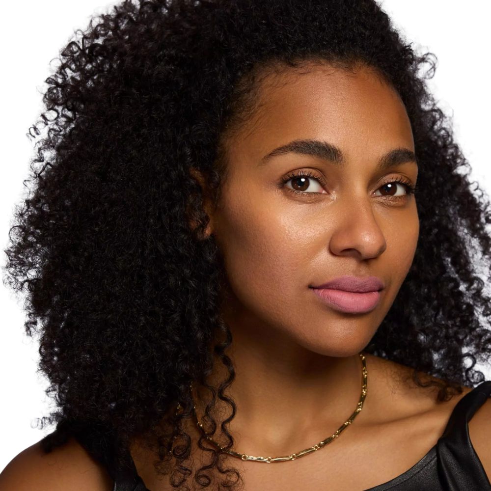 Portrait d'une femme aux cheveux noirs bouclés et collier doré. Elle porte du rouge à lèvres rose.