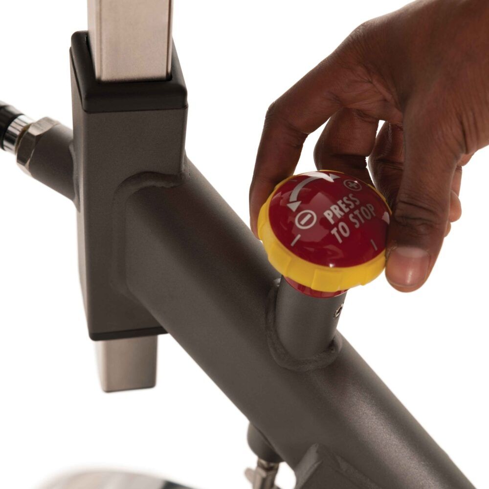 Close-up van de indoor cycle. Rode stopknop met gele ring. Opschrift: PRESS TO STOP. Grijs frame.